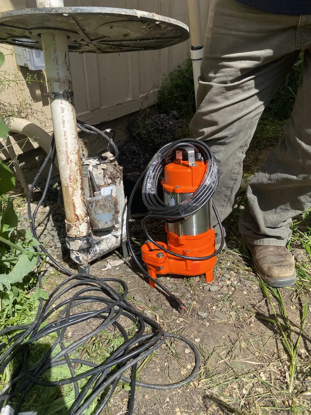An old, rusty well pump stands next to a new, orange replacement pump, with a person in work boots nearby.